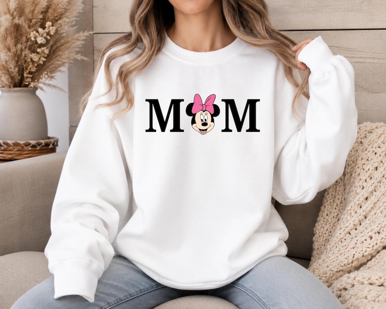 May include: White sweatshirt with the word "MOM" in black block letters. The "O" is replaced with a Minnie Mouse graphic wearing a pink bow. The sweatshirt has long sleeves and a crew neck.