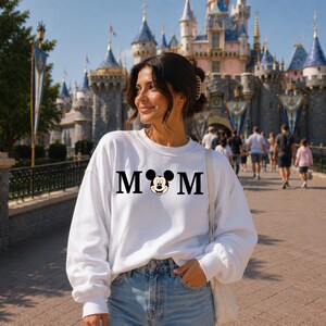 May include: A white sweatshirt with the word "MOM" in black block letters, with a Mickey Mouse head replacing the "O". The sweatshirt is worn by a person in front of a castle.