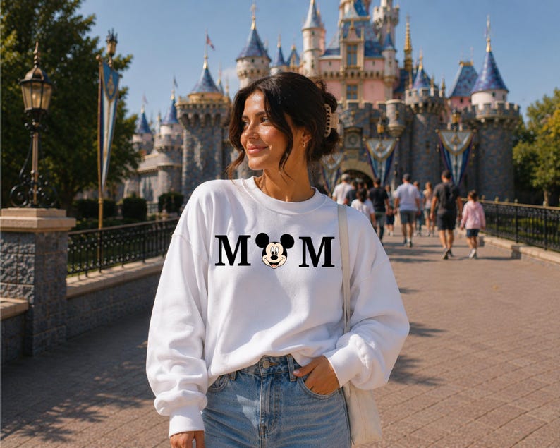 May include: A white sweatshirt with the word "MOM" in black block letters, with a Mickey Mouse head replacing the "O". The sweatshirt is worn by a person in front of a castle.