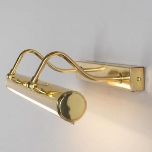 Peut inclure: Une applique murale pour tableau en métal doré poli, composée d'une base rectangulaire, de deux bras de support incurvés et d'une tête de lampe cylindrique. Le design comprend des articulations réglables pour orienter la lumière.
