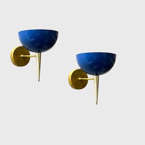 Può includere: Una coppia di applique da parete con paralumi a forma di ciotola blu intenso fissati a supporti circolari color oro. Ogni lampada presenta una sottile punta color oro che si estende verso il basso dalla base della ciotola blu.
