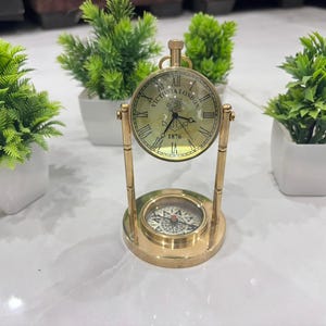 May include: A brass desk clock and compass, vintage in style. The clock face displays "VICTORIA LONDON 1876" in black Roman numerals. A compass is at the base. The clock is supported by a brass stand. Green plants in white pots are in the background.