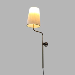 Może przedstawiać: Lampa ścienna z beżowym abażurem z tkaniny i mosiężnym metalowym ramieniem. Klosz ma kształt stożka i emituje ciepłe światło. Lampa jest przymocowana do ściany za pomocą okrągłej metalowej płyty i ma czarny przewód zasilający.