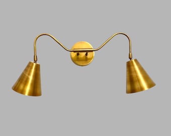 May include: A brass-toned wall sconce with two conical shades. The fixture has a circular base and curved arms extending to the shades. The shades are angled downward, and the overall design is minimalist and modern.