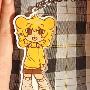 May include: An acrylic keychain featuring a cartoon character with yellow hair and bear-like ears, wearing a yellow long-sleeved shirt, brown shorts, and light-colored boots. The charm is attached to a silver-toned metal star-shaped clasp.