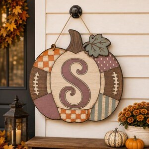 May include: A decorative wooden pumpkin wall hanging featuring a large purple letter S in the center. The pumpkin is divided into segments with various patterns including orange checkers, white polka dots, stripes, and football lace designs.