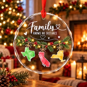Op de afbeelding: Een rond ornament van helder glas met een gefacetteerde rand, voorzien van de tekst Family KNOWS NO DISTANCE. Onder de tekst bevindt zich een groene dennentak met rode bessen, waaraan drie aquarelachtige kaarten van staten hangen: groen Texas, rood Californië en geel New York.