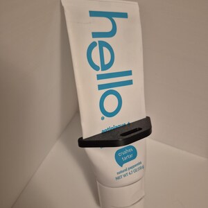 Pode incluir: Um tubo branco de pasta de dentes "hello" de hortel&atilde;-pimenta natural, preso em um suporte preto. O tubo tem texto azul e uma bolha azul que diz "crushes tartar". Peso l&iacute;quido 133g.