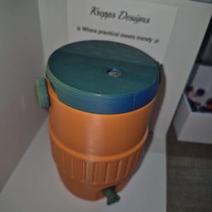May include: An orange and teal water cooler with a teal lid and a spigot. The cooler has a cylindrical shape with vertical ridges and a handle on the side. The text "Kropps Designs" is visible on a white sign.