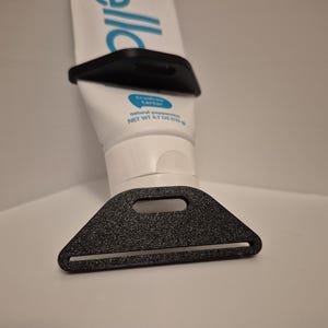 Pode incluir: Um espremedor e suporte de tubo de pasta de dentes de pl&aacute;stico preto. O espremedor tem formato triangular com uma ranhura para a passagem do tubo. O suporte tem formato retangular. O tubo de pasta de dentes &eacute; branco com texto azul.