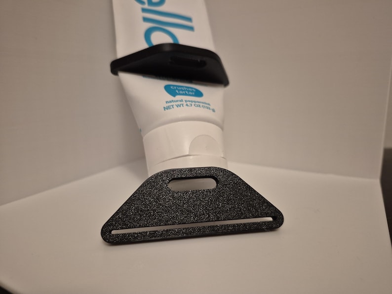 Pode incluir: Um espremedor e suporte de tubo de pasta de dentes de pl&aacute;stico preto. O espremedor tem formato triangular com uma ranhura para a passagem do tubo. O suporte tem formato retangular. O tubo de pasta de dentes &eacute; branco com texto azul.