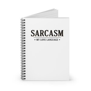 May include: A white, spiral-bound notebook with the word "SARCASM" in bold black letters. Underneath, the text reads "MY LOVE LANGUAGE". The notebook is open, showing blank white pages. The spiral binding is black.