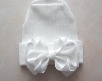 May include: A solid white knit beanie hat featuring a large, centered, multi-layered satin ribbon bow in the same white color. The hat has a folded cuff at the bottom, and the entire piece is constructed from a consistent, plain white textile.