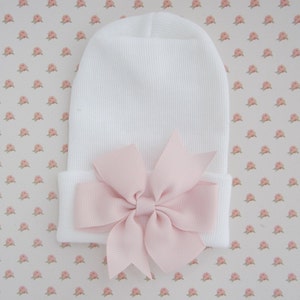 May include: A white knit beanie hat featuring a folded cuff and a large, light pink grosgrain ribbon bow centered on the front. The hat is constructed from a textured, ribbed fabric and displays a simple, clean design with a soft pastel accent.