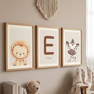 Könnte beinhalten: Drei gerahmte Kunstdrucke für das Kinderzimmer. Einer zeigt einen Löwen, ein weiterer den Buchstaben "E" mit dem Namen "Elias" und der dritte ein Zebra. Die Drucke haben eine neutrale Farbpalette mit Holzrahmen.