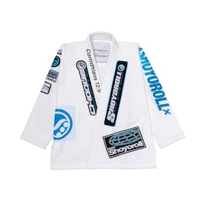 May include: White martial arts uniform with various patches and text. The uniform displays the text "SHOYOROLL" in blue on the sleeves and chest. Additional patches include a globe design and the text "Corinthians 12:9".
