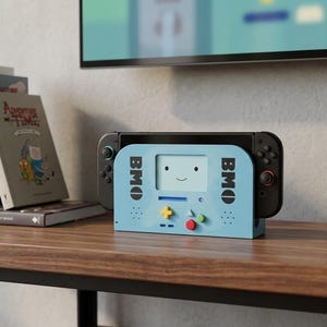 Op de afbeelding: Een lichtblauw dockingstation voor een spelcomputer met het personage BMO uit Adventure Time. Het dock heeft een schermafbeelding met een lachend gezicht, zwarte tekst met BMO aan beide kanten en kleurrijke ronde knoppen in geel, groen en rood.