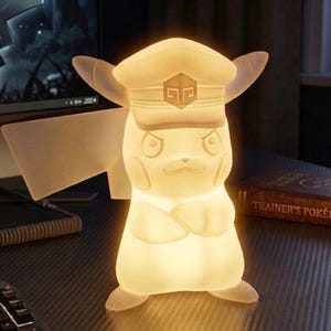 May include: A glowing Pikachu-shaped lamp wearing a hat with a badge. The lamp is on a desk with a computer monitor and a book titled "Trainer's Pokédex". The lamp is a soft, warm white color.