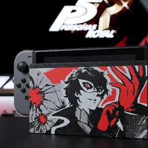 May include: A grey video game console dock featuring a custom graphic print of a character wearing a black mask and red gloves, with shattered glass patterns in red and black. The dock is attached to a grey handheld gaming console with controllers.