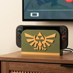 May include: A gaming console dock featuring an olive green front panel with a raised gold-colored Triforce emblem. The dock holds a black handheld gaming device with black controllers attached, positioned on a wooden surface near a small potted succulent.