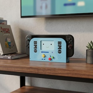 May include: A light blue gaming console dock featuring a BMO character design with a smiling face, black text, and colorful buttons. The dock holds a black handheld gaming device and is positioned on a wooden surface next to books and a small plant.