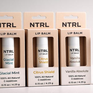 May include: Lip balm products from NTRL, featuring Glacial Mint, Citrus Shield, and Vanilla Absolute flavors. Each product is in a white tube and packaged in a tan cardboard box. The boxes and tubes display the brand name and product information.