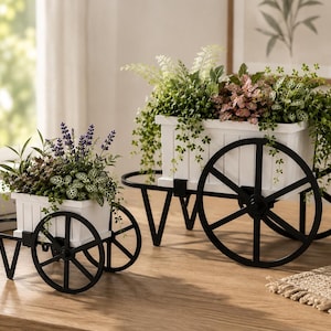 May include: Two decorative white wooden planter carts with black metal wheels. The carts are filled with various green and purple plants. One cart is smaller than the other. The carts are on a wooden table.
