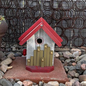 May include: A handcrafted birdhouse with a red roof and base, and a gray and white body. The birdhouse features a circular entry hole and decorative gold-colored wooden accents. It is set on a red brick and surrounded by rocks.