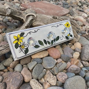 May include: A rectangular, light gray wooden wall hook with three crystal knobs. The hook is decorated with yellow sunflowers, green leaves, and a black vine design. The background is a bed of rocks and driftwood.