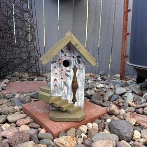 May include: A decorative birdhouse with a distressed white and gray finish, adorned with floral accents in pink and brown. It features a small staircase and a bronze-colored handle. The birdhouse is set on a red brick base, surrounded by rocks.