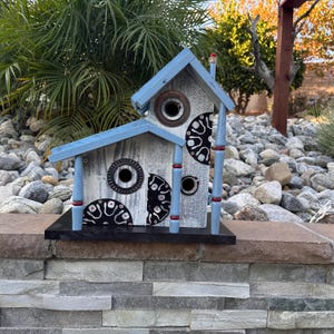 May include: A decorative birdhouse with a light blue roof and accents. The birdhouse is painted white with black and white circular designs and has a black base. The birdhouse has a chimney and is made of wood.