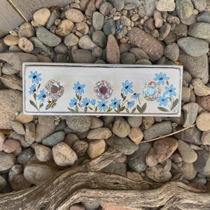 May include: A rectangular, white wooden wall hook with three crystal knobs. The board is decorated with blue flowers, green leaves, and a small bee. The knobs are clear and red. The artwork is on a bed of rocks.