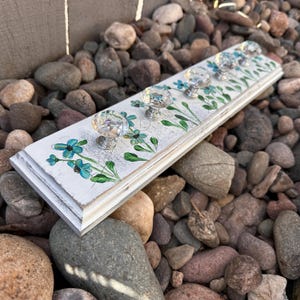 May include: A white wooden wall hook with five clear crystal knobs. The board is decorated with hand-painted turquoise flowers and green leaves. The edges of the board are distressed, giving it a rustic look. The wall hook is resting on a bed of rocks.
