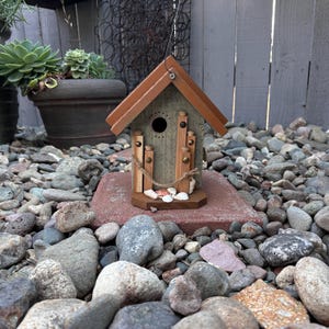 May include: A decorative birdhouse with a brown roof and a gray body, accented with wooden dowels and a small rope. The birdhouse sits on a red brick, surrounded by rocks and greenery, creating a rustic outdoor aesthetic.