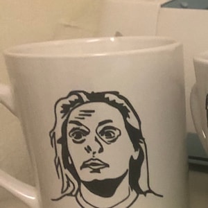 May include: White ceramic mug with a black and white illustration of a person's face.