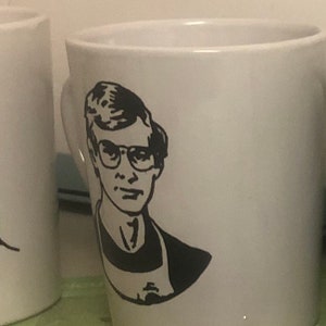 May include: White ceramic mug with a black and white illustration of a person wearing glasses. The person has short, dark hair and is wearing a dark shirt.