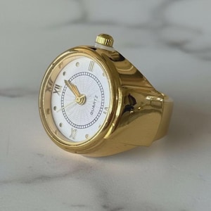 Op de afbeelding: Een goudkleurige ringhorloge met een witte wijzerplaat en zwarte Romeinse cijfers. Het horloge heeft een quartz uurwerk en een goudkleurige kroon. De ring is een uniek sieraad.