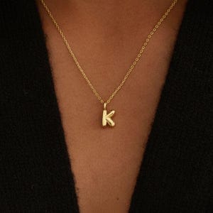 Può includere: Una collana dorata con una catena sottile e un ciondolo a forma di lettera "K". Il ciondolo è piccolo e centrato, su uno sfondo scuro. Un gioiello semplice ed elegante.