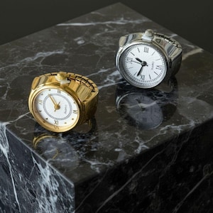 Può includere: Due orologi ad anello, uno dorato e uno argentato, con quadranti bianchi e numeri romani. Gli orologi sono su una superficie di marmo nero, che riflette le loro immagini. L'orologio dorato è a sinistra e quello argentato a destra.