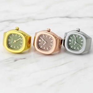 Può includere: Tre anelli orologio in oro, oro rosa e argento. Ogni anello orologio ha un quadrante quadrato con un quadrante colorato e un cinturino abbinato. I quadranti hanno linee verticali e le lancette indicano orari diversi.