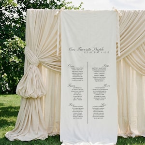 Puede incluir: Un plano de asientos de boda de color crema con el texto "Our Favorite People, Please Take a Seat". El gráfico enumera los nombres bajo los encabezados "One" a "Six". El telón de fondo está cubierto con tela color crema.