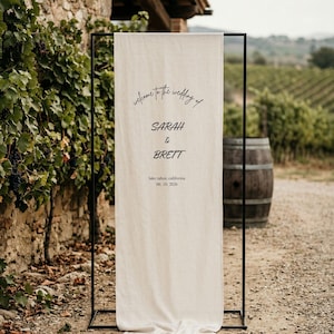 Puede incluir: Un cartel de bienvenida de boda color crema con texto negro que dice "welcome to the wedding of SARAH & BRETT" y la fecha "06.10.2026". El letrero se muestra en un marco de metal negro, al aire libre con un telón de fondo de viñedos.