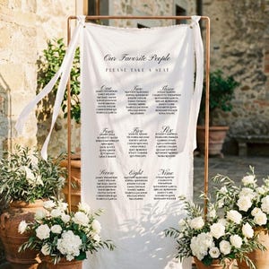 Puede incluir: Un plano de asientos de boda en tela blanca, enmarcado por una estructura de cobre. El texto "Our Favorite People" y "Please Take a Seat" es visible. Flores blancas en macetas de terracota flanquean la tabla, creando un entorno exterior elegante.