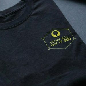 Op de afbeelding: Zwart t-shirt met een geel geometrisch ontwerp en de tekst "ESCAPE WILL MAKE ME GOD". Het shirt is op een donkere ondergrond gevouwen. Een toetsenbord, muis en klein beeldje staan op de achtergrond.