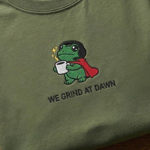 Puede incluir: Camiseta verde oliva con un bordado de una rana con capa y taza de café. El texto "WE GRIND AT DAWN" está debajo. La camiseta es de algodón grueso.