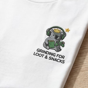 Peut inclure: T-shirt blanc avec un motif brodé d'un koala portant un casque et une couronne, tenant une manette de jeu et une boîte verte. Le texte "GRINDING FOR LOOT & SNACKS" est en dessous. Une tasse à café avec un cœur pixélisé est également visible.
