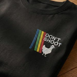 Op de afbeelding: Zwart T-shirt met ronde hals en geborduurd ontwerp. Het ontwerp toont een regenboog van verticale strepen, de woorden "DON'T SHOOT" en een witte kip met hoed.