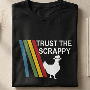 Könnte beinhalten: Schwarzes T-Shirt mit dem Text "TRUST THE SCRAPPY" und einem weißen Huhn mit Hut. Das Design beinhaltet einen diagonalen Streifen aus Blau, Grün, Gelb und Rot. Ein humorvolles Grafik-T-Shirt.