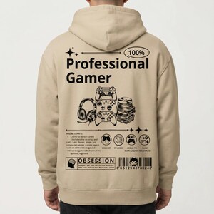 May include: A beige hoodie featuring the text "100% Professional Gamer" and gaming-related graphics. The design includes headphones, controllers, and a stack of discs. The back of the hoodie also has additional text and a barcode.