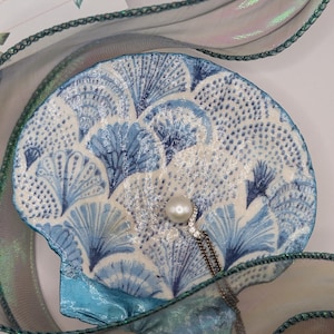 May include: A decorative seashell-shaped dish with a blue and white fan pattern. A pearl pendant necklace rests on the dish. Iridescent ribbon in shades of green and blue frames the seashell, creating a coastal theme.